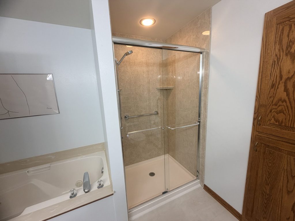 Walk-in shower with beige stone-look wall panels, a sliding glass door, handheld shower and grab bar, installed in Greendale, WI by TightSeal Exteriors & Baths