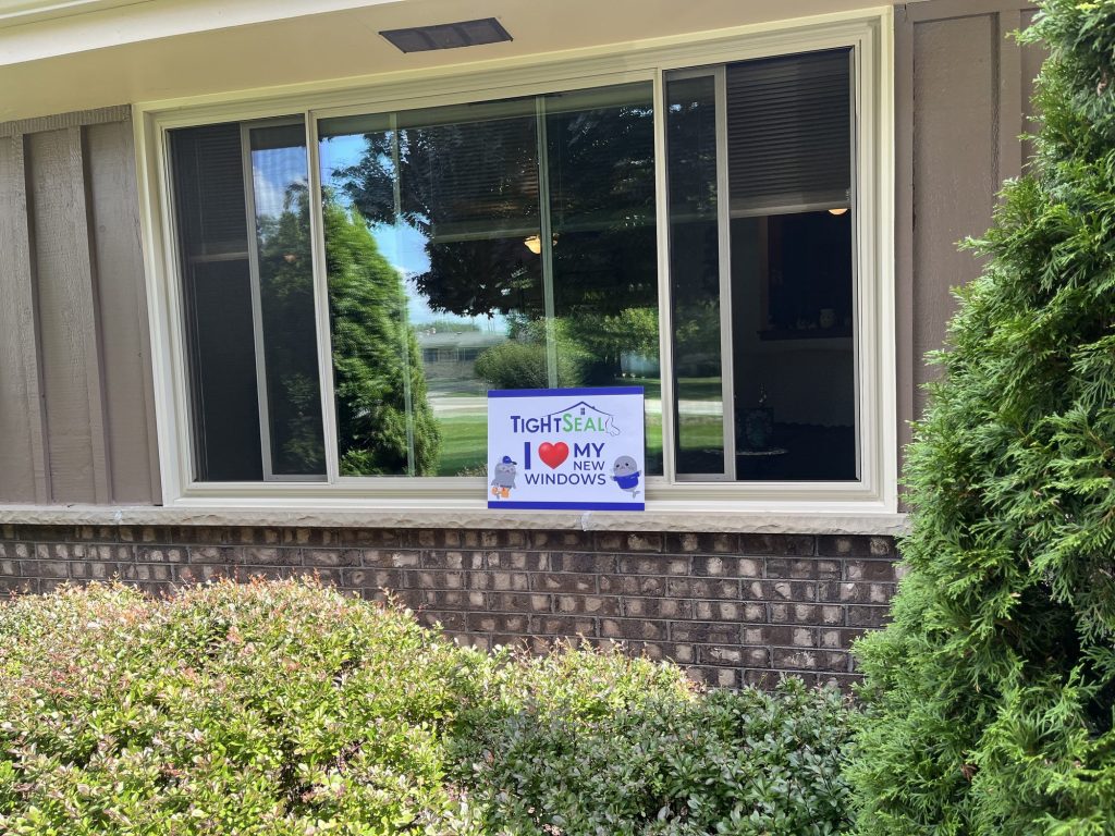 Large picture window with sliding side sashes and a tan frame, installed on a brick and board-and-batten Muskego, WI home by TightSeal Exteriors & Baths
