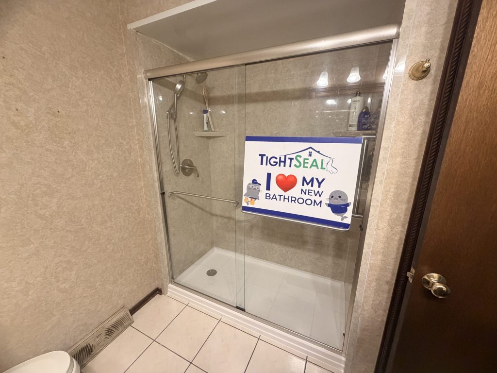 Walk-in shower with beige stone-look wall panels, a brushed-nickel sliding glass door and handheld shower, installed in Muskego, WI by TightSeal Exteriors & Baths