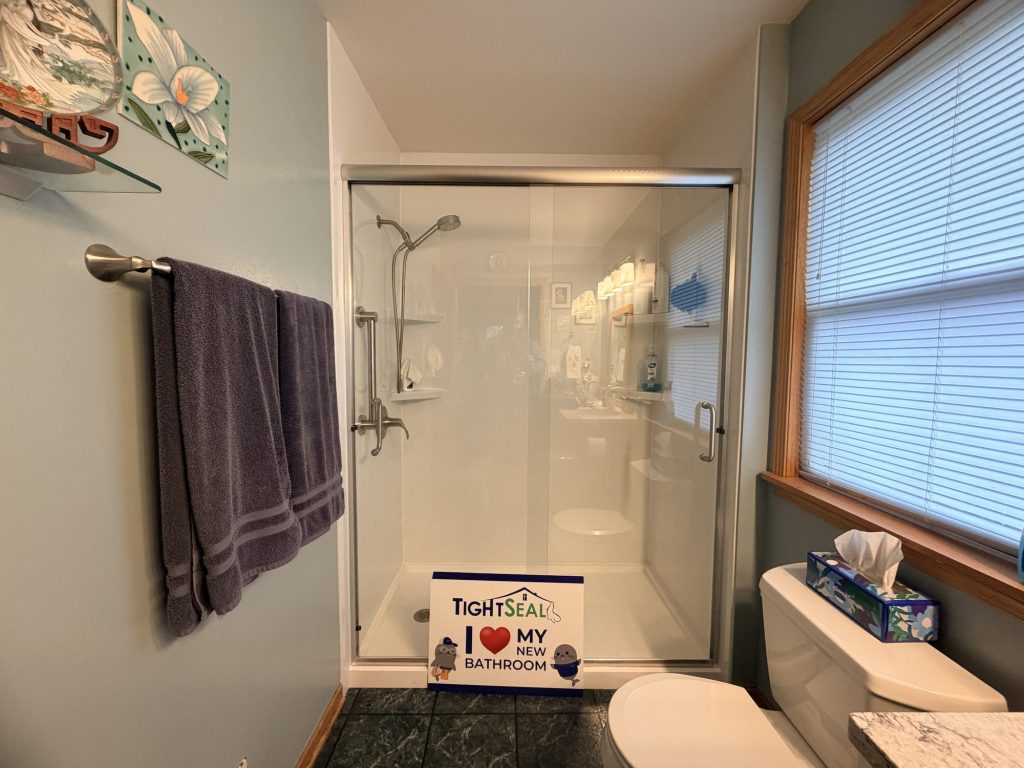Walk-in shower with white wall panels, a brushed-nickel sliding glass door, handheld shower and grab bar, installed in Oconomowoc, WI by TightSeal Exteriors & Baths
